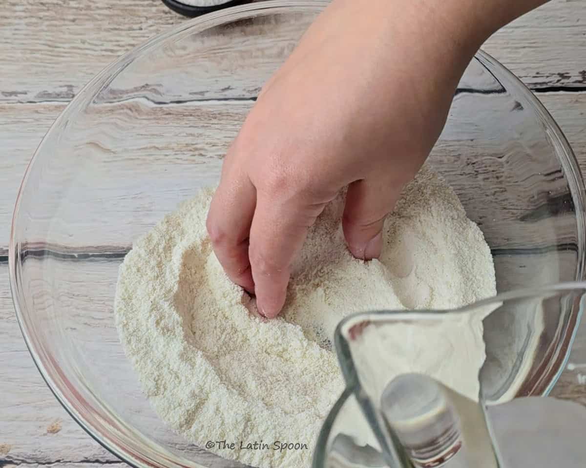 A hand touches the corn dough inside a large glass bowl as water is poured from a glass pitcher. In the background, there’s a small bowl with salt and two tomatoes.