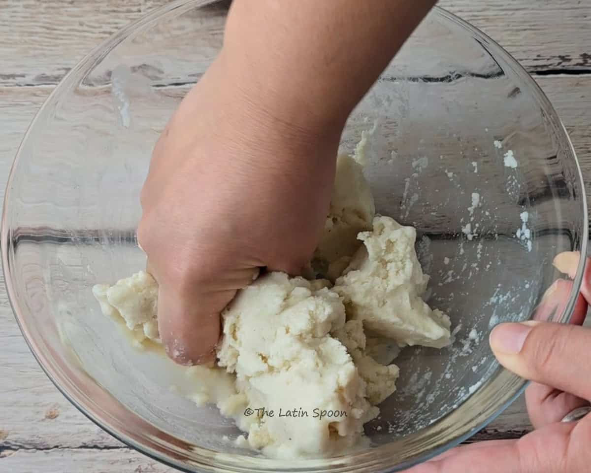 A hand mixes the corn dough inside a glass bowl, with a small bowl of salt in the background and tomatoes in the foreground.