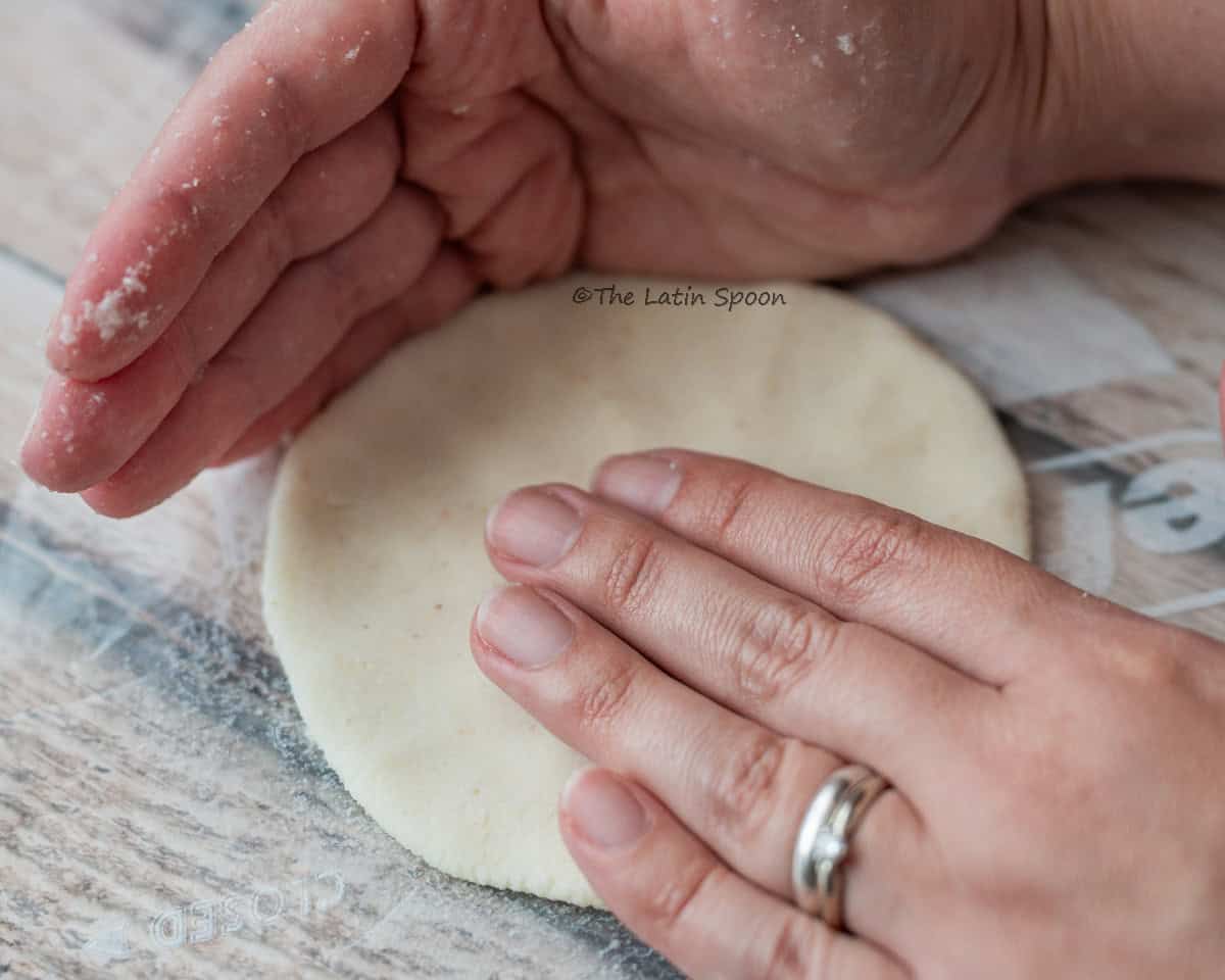 The right hand presses the dough into a tortilla shape over a circular piece of plastic, while the left hand forms a “C” shape to shape the edges.
