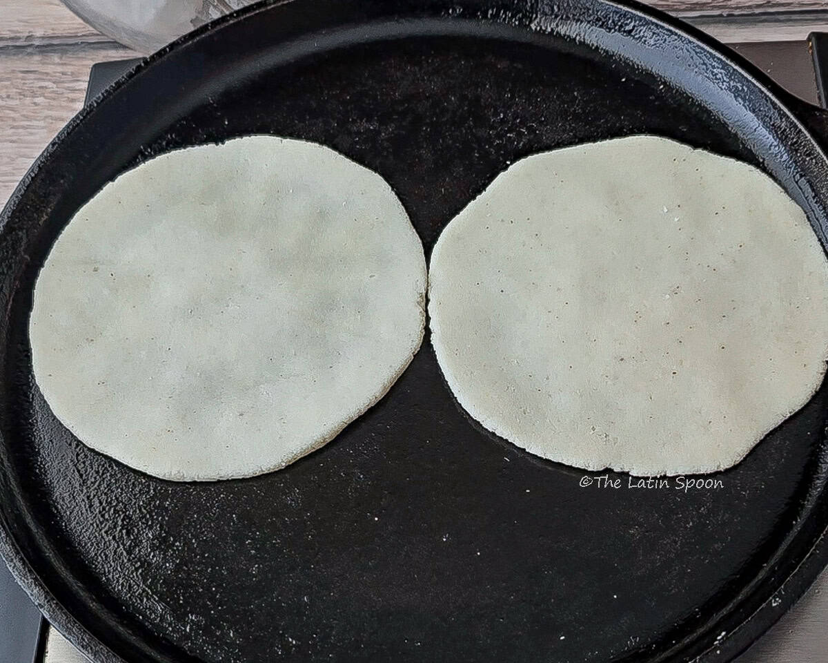 A comal on a stovetop cooking two tortillas, with a clear bowl holding an oiled napkin visible in the background.
