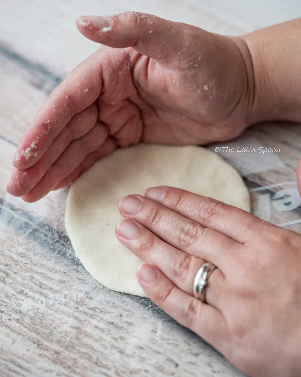 The right hand presses the dough into a tortilla shape over a circular piece of plastic, while the left hand forms a “C” shape to shape the edges.