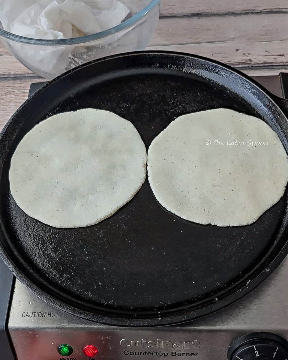 A comal on a stovetop cooking two tortillas, with a clear bowl holding an oiled napkin visible in the background.