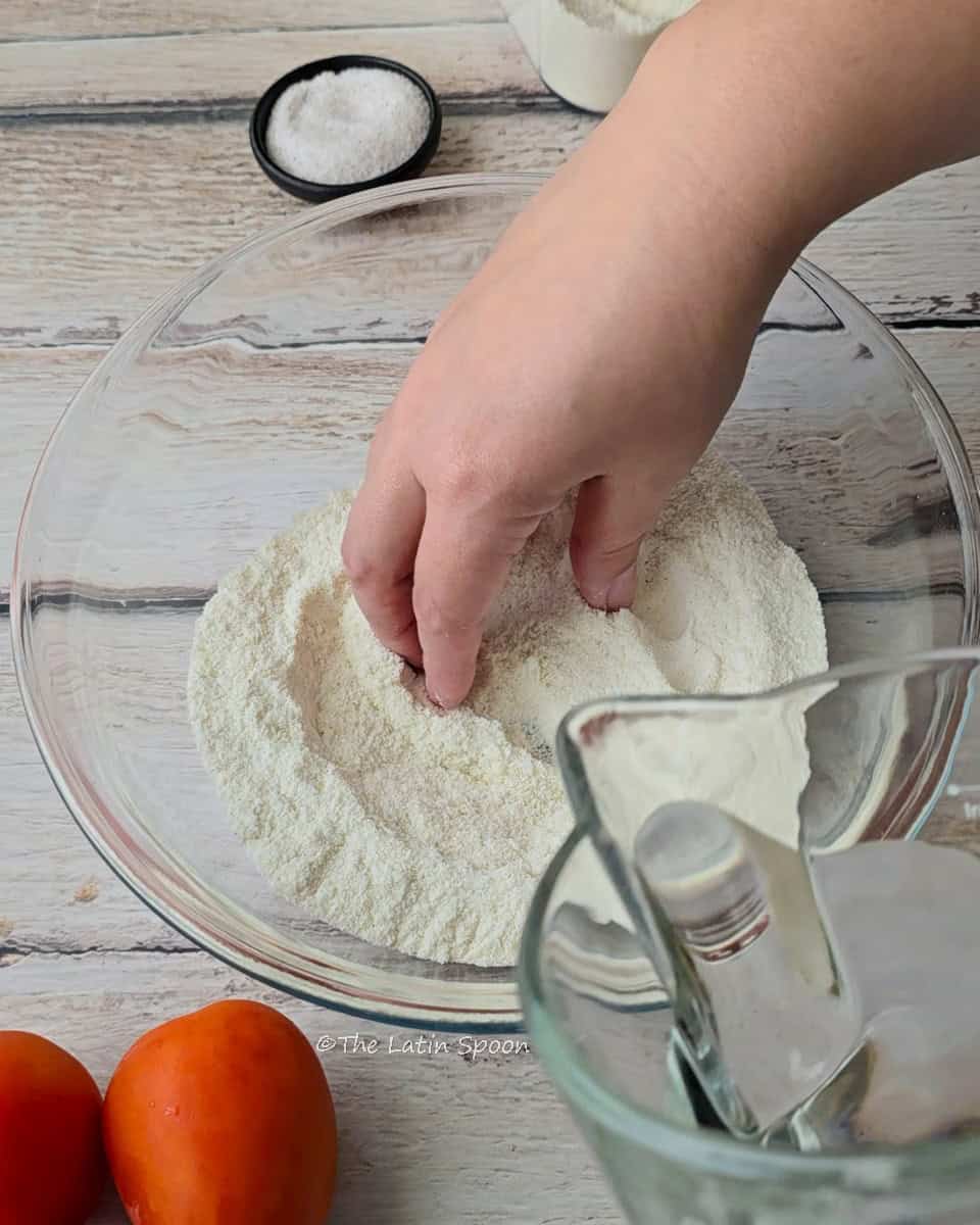 A hand touches the corn dough inside a large glass bowl as water is poured from a glass pitcher. In the background, there’s a small bowl with salt and two tomatoes.