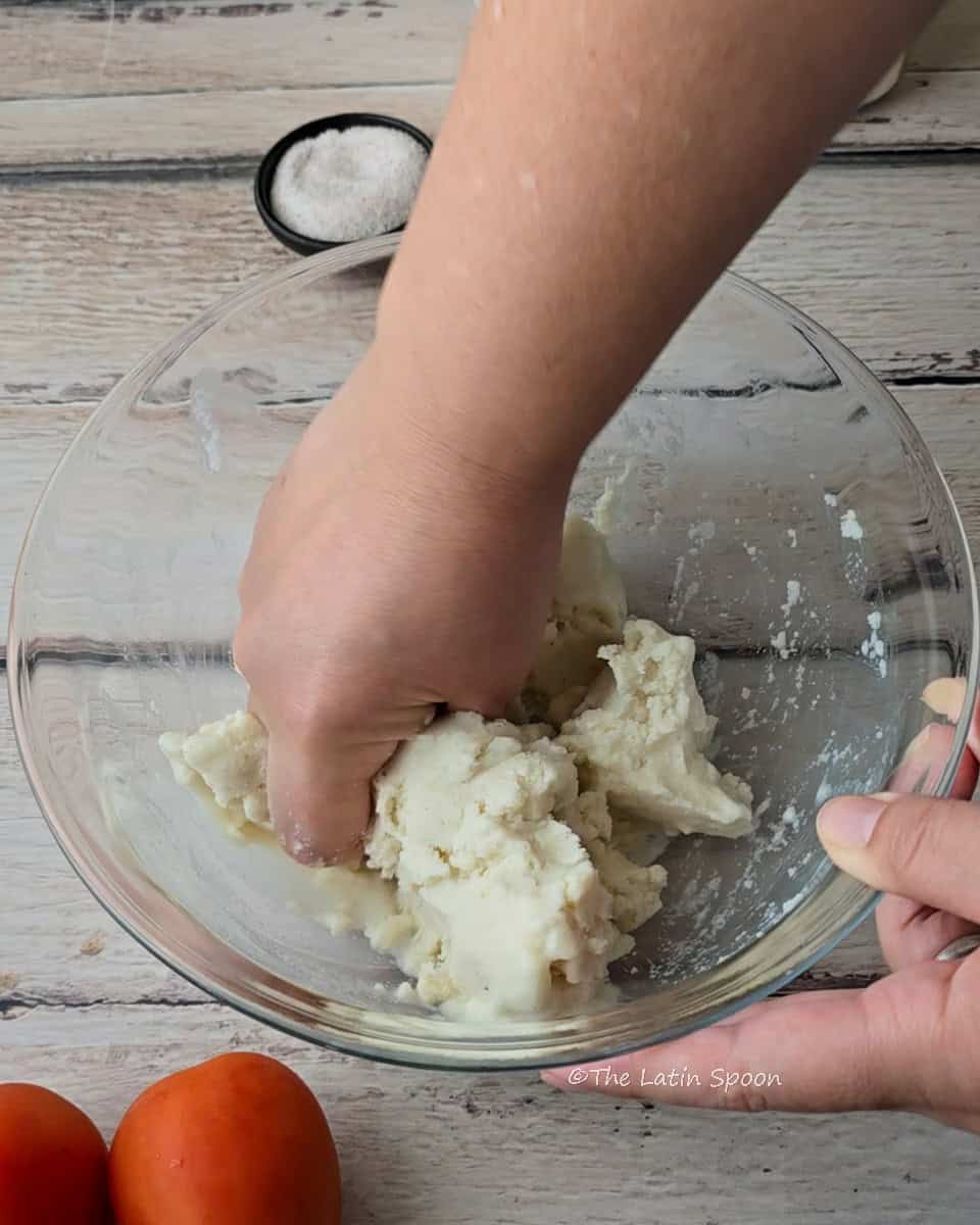 A hand mixes the corn dough inside a glass bowl, with a small bowl of salt in the background and tomatoes in the foreground.