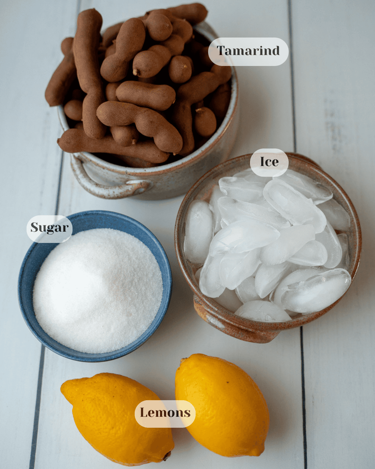 Top view of small bowls containing tamarind pods, ice cubes, and sugar, with two lemons placed next to them.