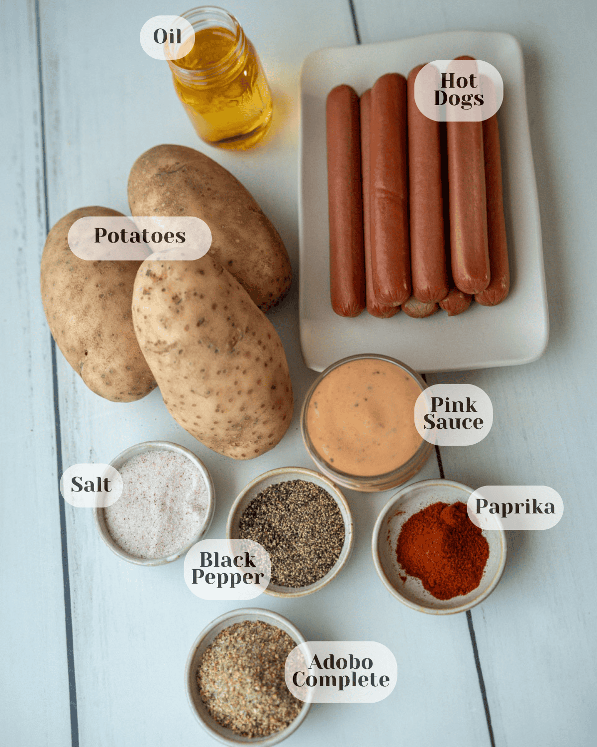 A small cup of olive oil, a plate with sausages and potatoes, and seasonings including salt, pink sauce, black pepper, paprika, and all-purpose seasoning.