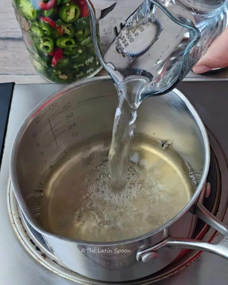 A small pot on the stove filled with vinegar as a measuring cup pours water into it, with a jar of jalapeños in the background.