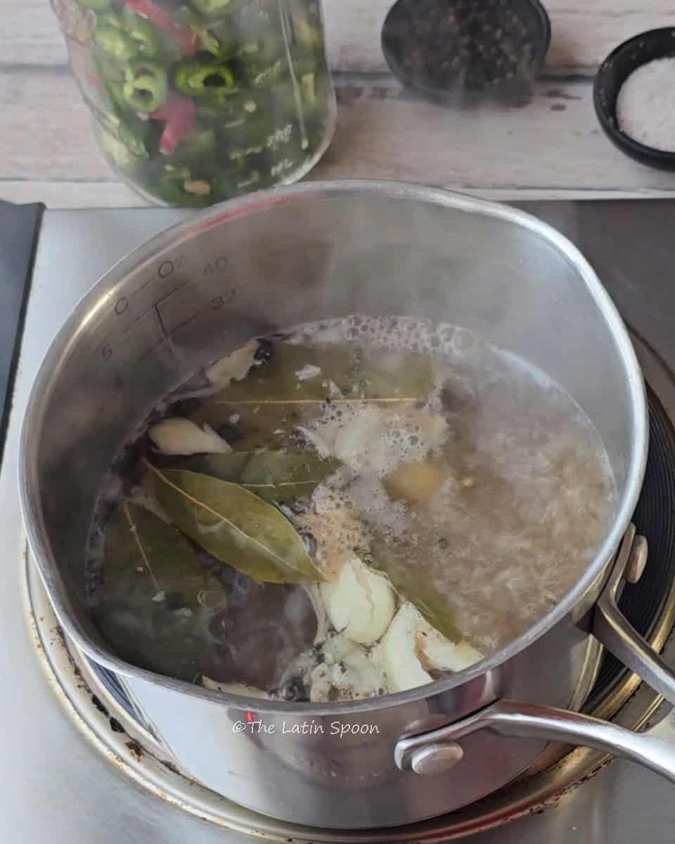 A small pot on the stove containing garlic, black peppercorns, bay leaves, vinegar, and water. Next to it, there’s a jar of jalapeños and a small cup with salt and pepper.