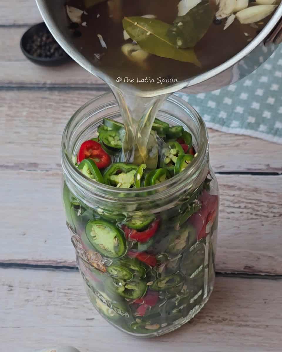 A jar filled with sliced jalapeños as a pot with hot brine and seasonings is poured over them.