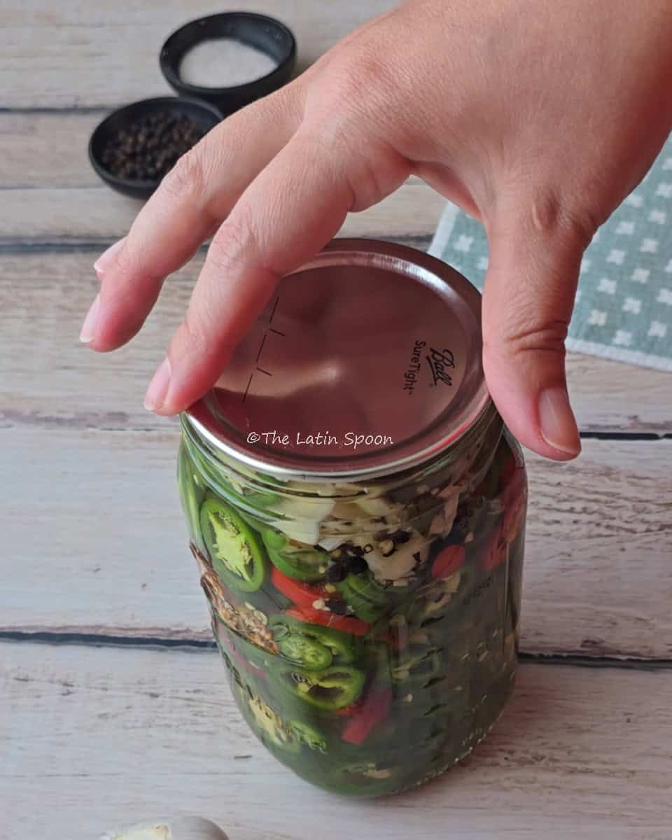 A hand placing the lid on a jar filled with jalapeños, with a small cup of salt and another of pepper in the background.