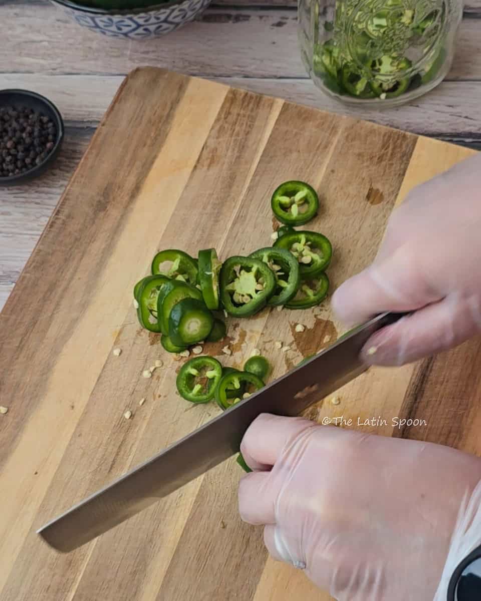 On a cutting board, the right hand holds a knife slicing jalapeños while the left hand steadies them. In the background, there’s a jar filled with sliced jalapeños and a small cup of whole peppercorns.