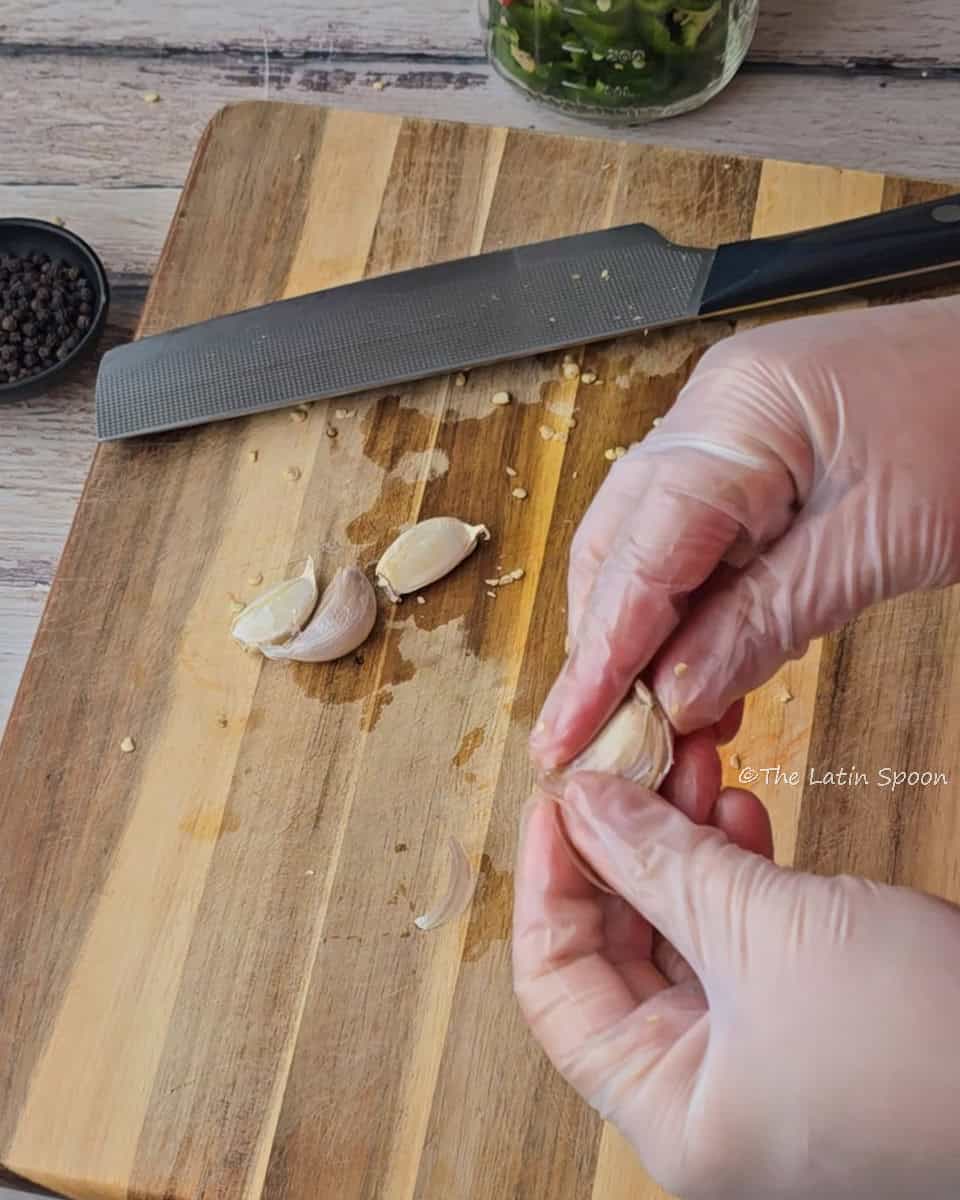 Two hands peel a garlic clove over a cutting board with three garlic cloves and a knife resting on it.