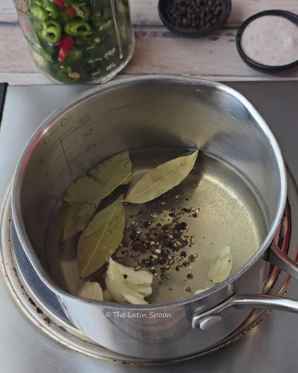 A small pot on the stove containing garlic, black peppercorns, bay leaves, vinegar, and water. Next to it, there’s a jar of jalapeños and a small cup with salt and pepper.