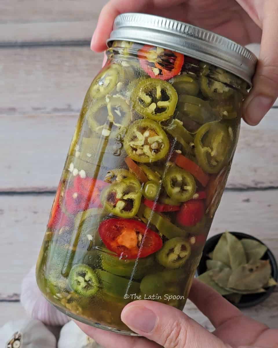 Sealed jar of homemade pickled jalapeños in brine, held by both hands, with bay leaves and garlic bulbs in the background.