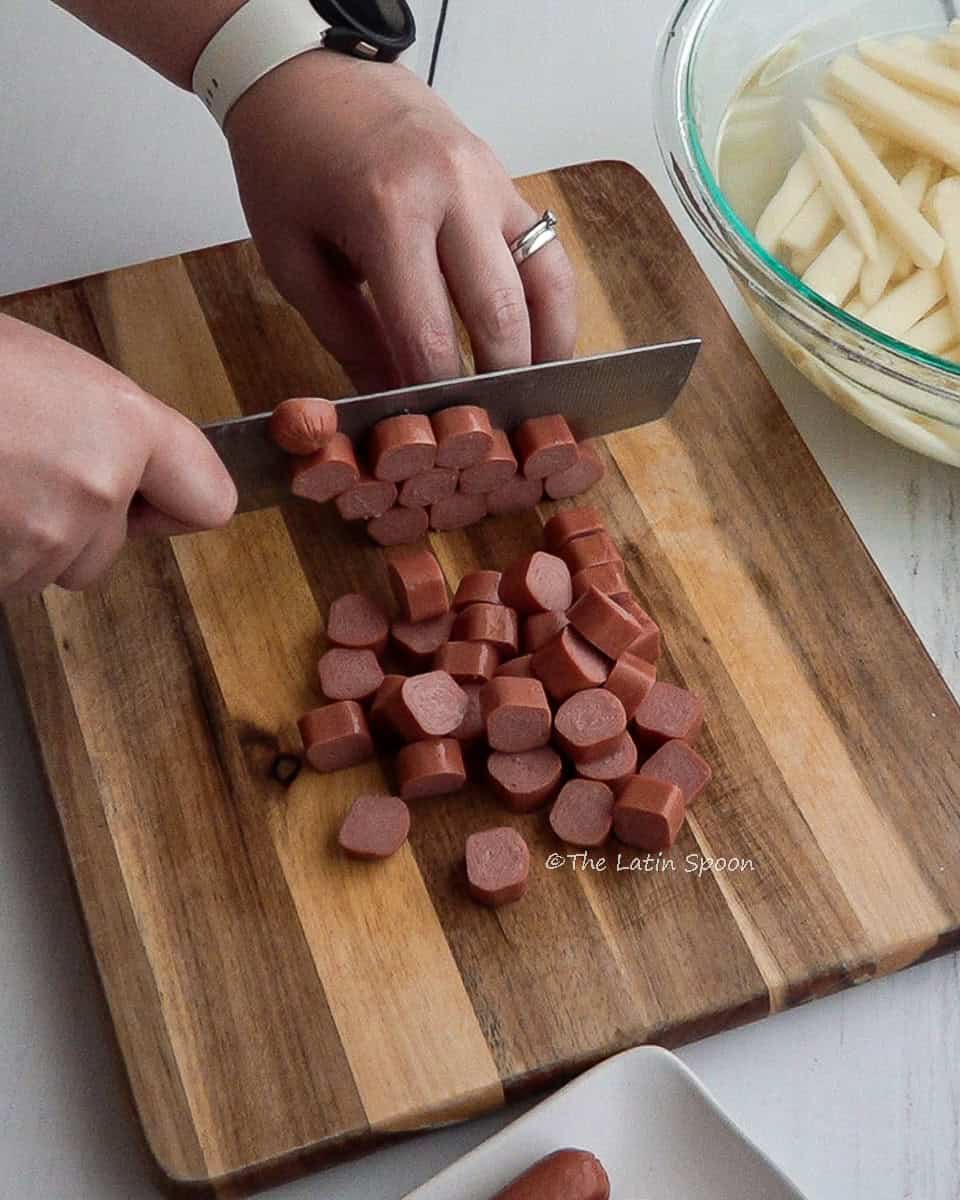 The left hand holds the sausages while the right hand cuts them into small pieces on a cutting board, with a bowl of cut potatoes in the background.