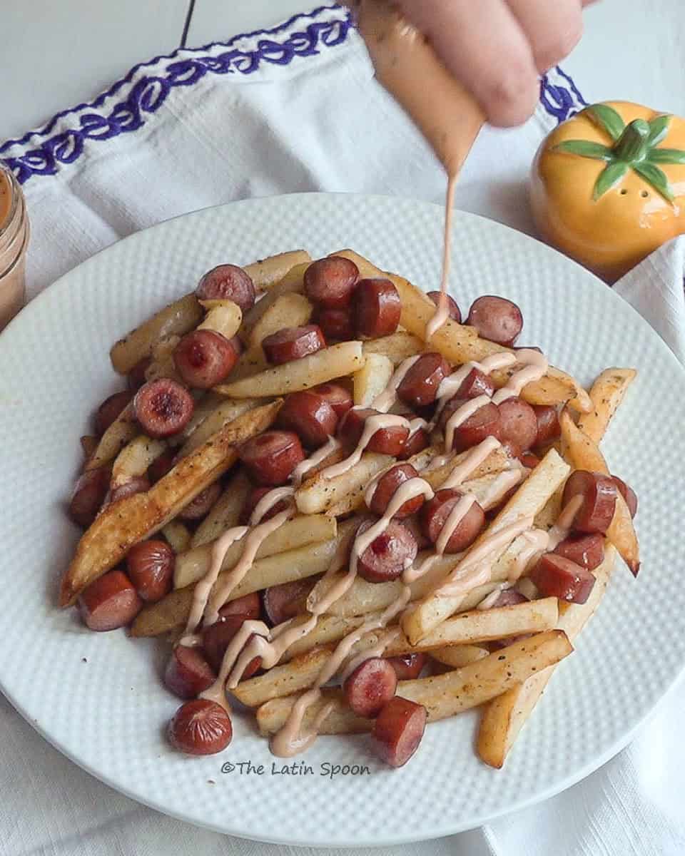 A plate of French fries and sliced sausage topped with pink sauce, with a tomato-shaped salt shaker in the background.