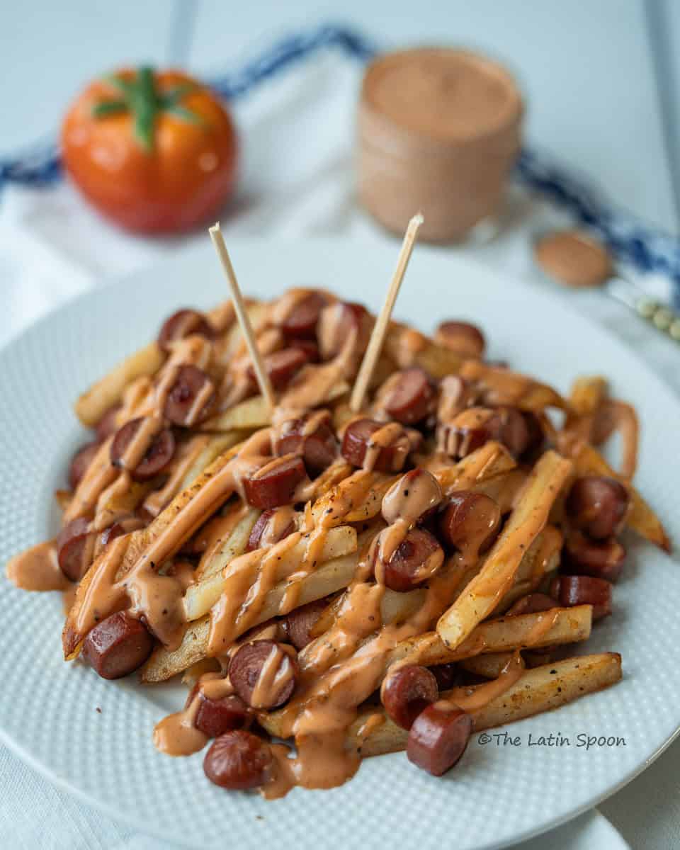 A plate of salchipapas with two wooden sticks on top, a tomato-shaped salt shaker, and a bottle of pink sauce in the background.