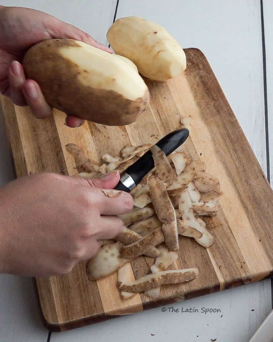A cutting board with potato peels falling as the left hand holds a potato and the right hand peels it, with a peeled potato in the background.
