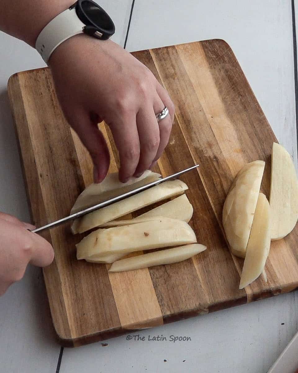 The right hand cuts a potato into strips with a knife while the left hand holds it steady on a cutting board.