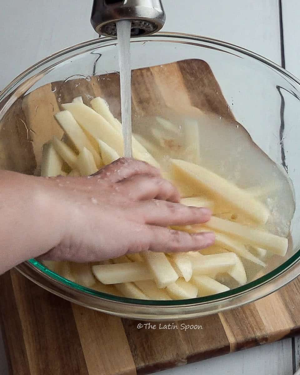 Cut potato strips in a large bowl set on a cutting board, rinsed with tap water while the right hand stirs them.