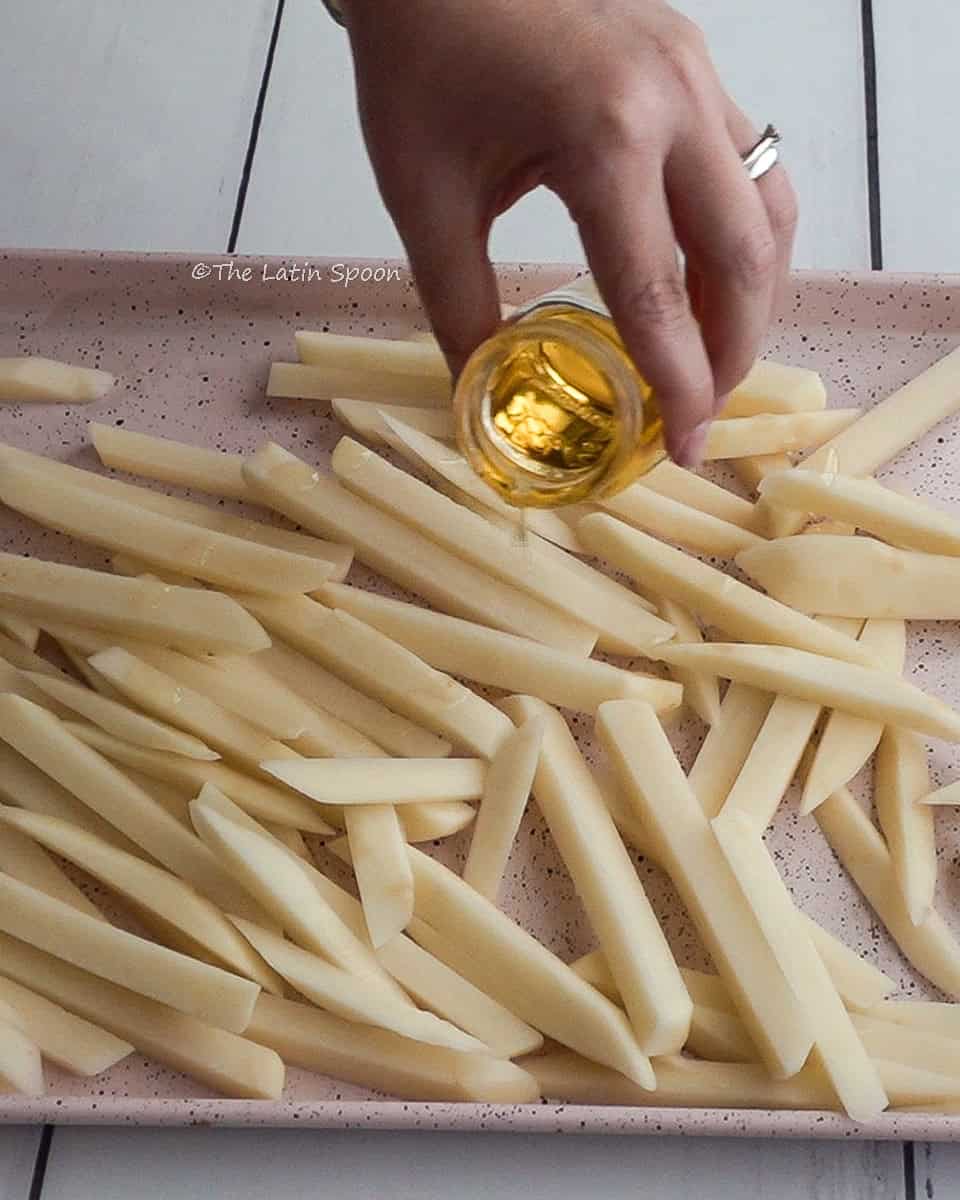 Cut potato strips spread on a tray while the right hand pours oil from a container over them.