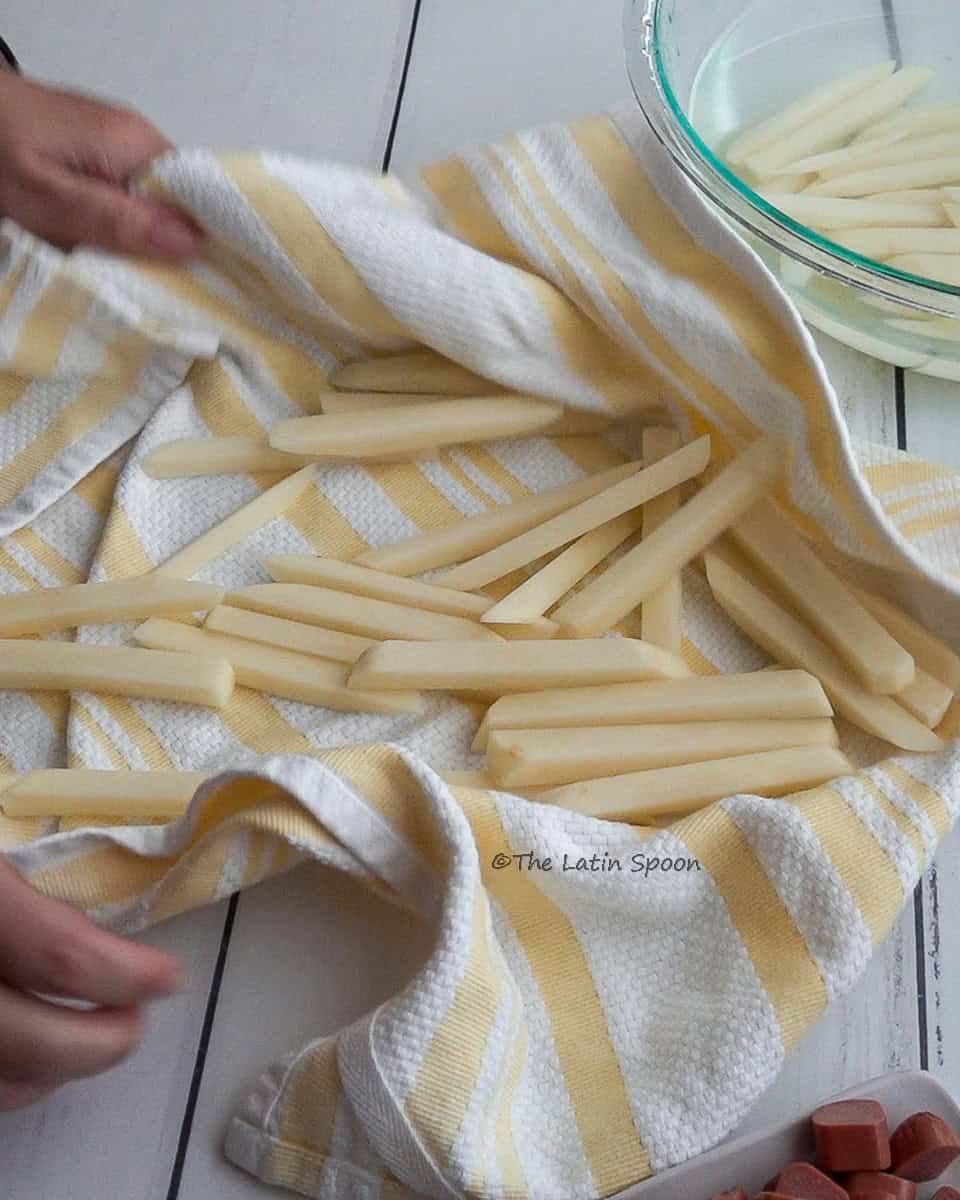 Hands hold a kitchen towel filled with potato strips, drying them completely. A bowl with more potatoes strips sits in the background.