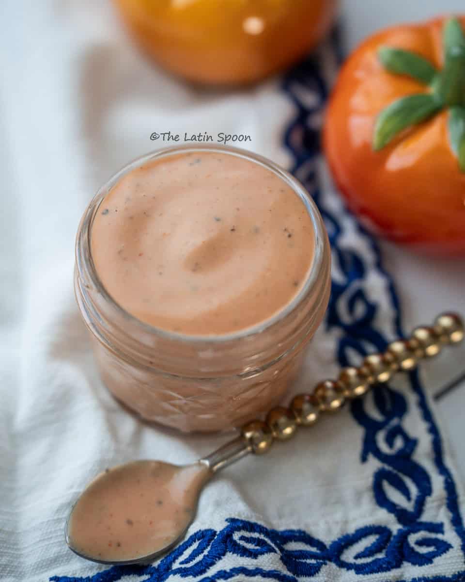 A small cup of creamy pink sauce sits on a decorative cloth, with a spoon nearby and tomato-shaped salt and pepper shakers behind it.
