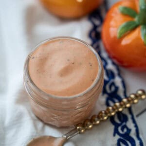 A small bowl of pink sauce on a decorative cloth, with a spoon and cherry tomatoes in the background.