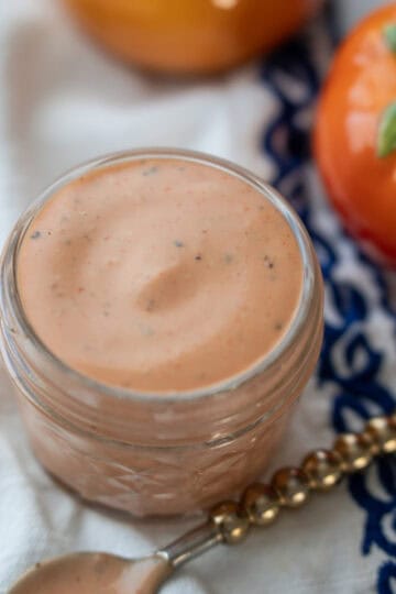A small bowl of pink sauce on a decorative cloth, with a spoon and cherry tomatoes in the background.
