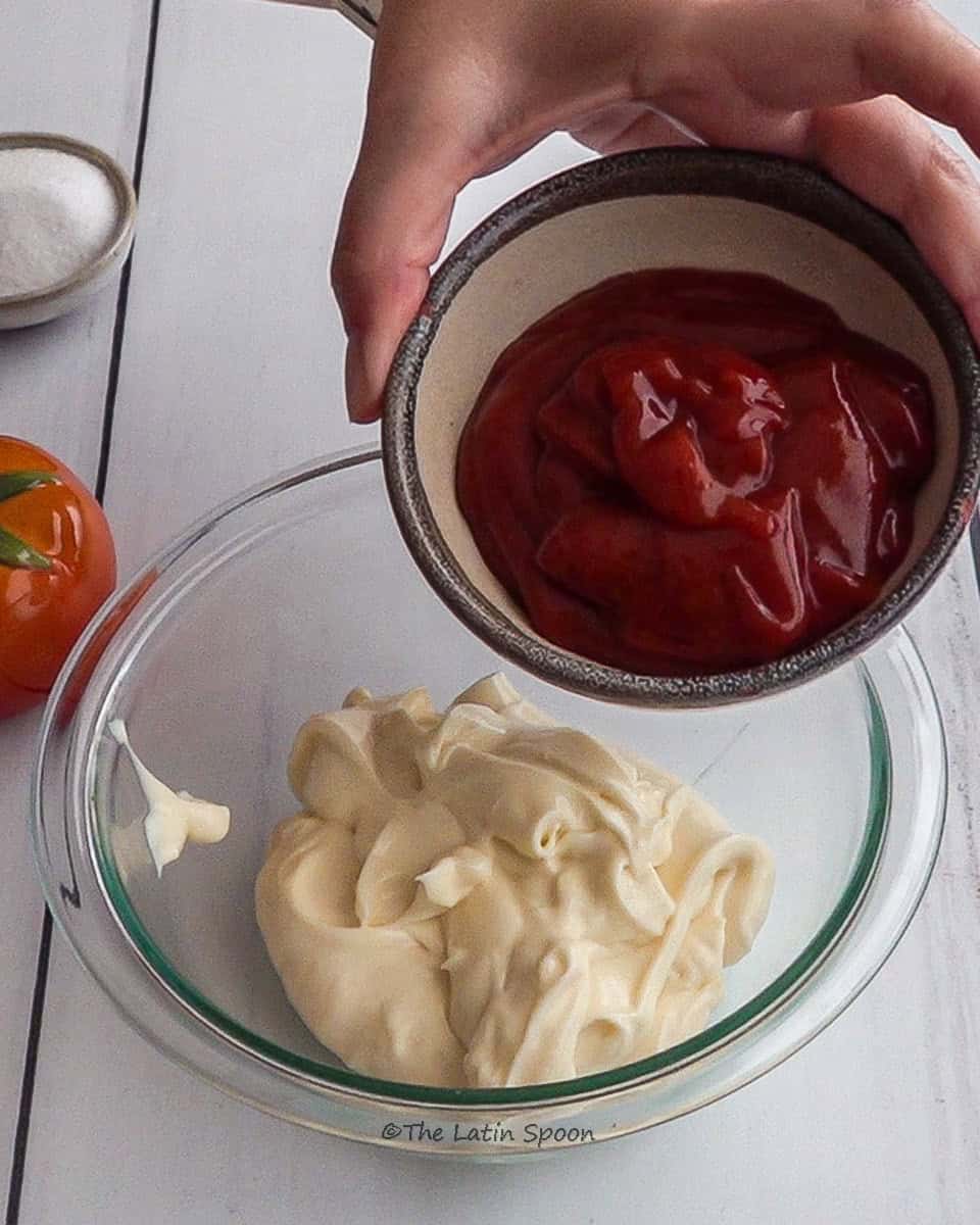 A left hand pouring tomato sauce from a small cup into a bowl of mayonnaise, with a small cup of sugar and a decorative tomato in the background.