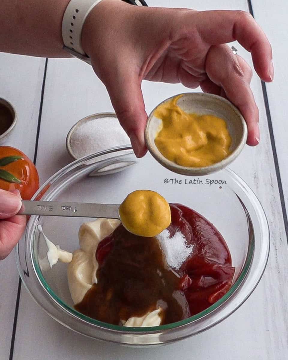 A left hand holding a small cup of mustard and a right hand holding a measuring spoon filled with mustard over a bowl containing mayonnaise, tomato sauce, Lizano sauce, and sugar.