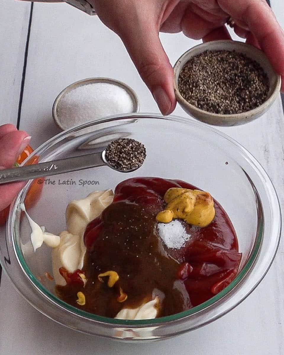 A bowl with mayonnaise, tomato sauce, Lizano sauce, mustard, and sugar, while the left hand holds a small cup of pepper and the right hand sprinkles it over the mixture.