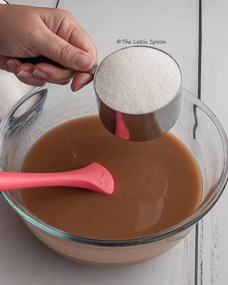 A left hand holds a measuring cup with sugar over a bowl containing tamarind pulp. A spatula rests inside the bowl.