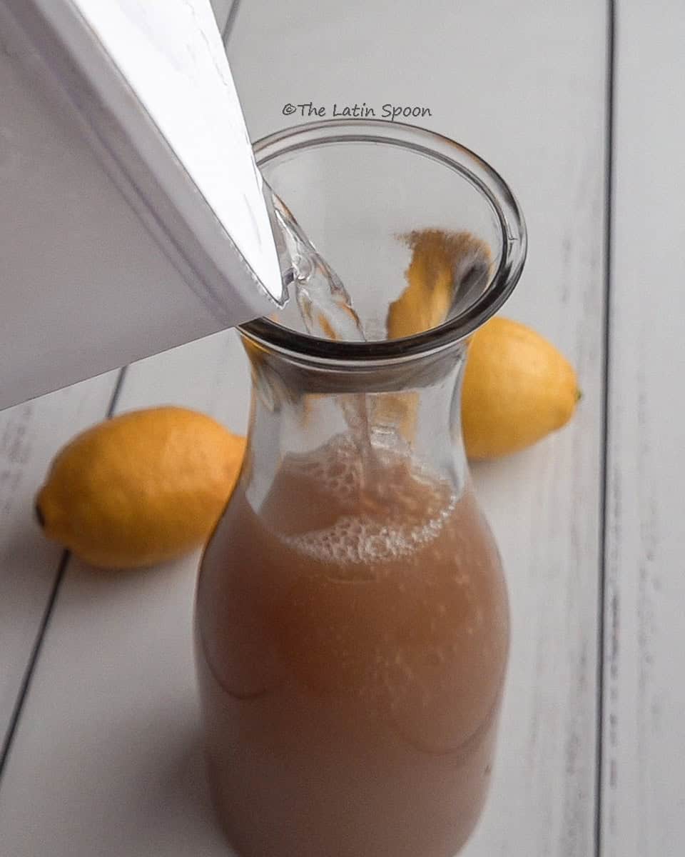 A pitcher filled with tamarind pulp mixture, while another pitcher pours water into it. In the background, two lemons are visible.