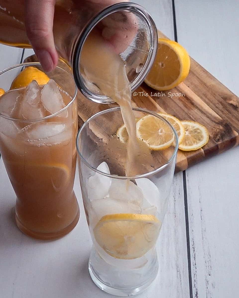 Two glasses: one filled with tamarind drink and ice, the other with ice and lemon slices while a pitcher pours in the tamarind drink. In the background, a wooden board holds lemon slices and half a cut lemon.