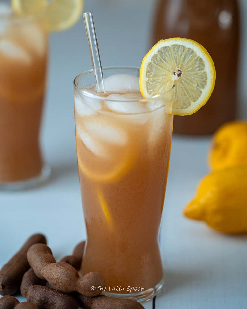 A glass filled with fresco de tamarindo, decorated with lemon slices on the rim and floating inside the drink. At the base, a few tamarind pods are arranged decoratively, their rustic brown shells adding texture. In the background, a couple of fresh lemons and two more glasses of the drink