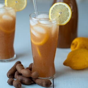 A glass of tamarind drink with yellow lemon slices on the rim and inside the glass. Decorative tamarind pods are placed at the base. In the background, there are a few lemons and two more glasses of the same drink.