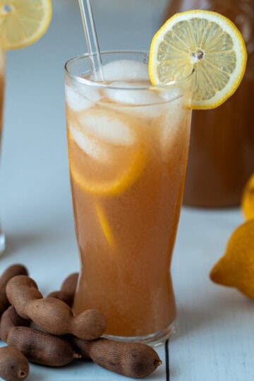 A glass of tamarind drink with yellow lemon slices on the rim and inside the glass. Decorative tamarind pods are placed at the base. In the background, there are a few lemons and two more glasses of the same drink.