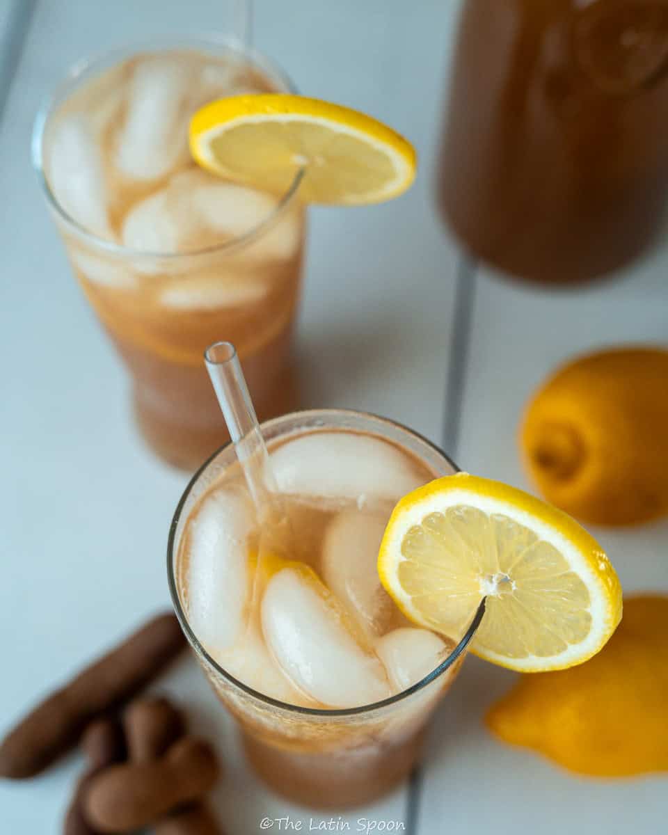 Top view of two glasses of tamarind drink decorated with lemon slices. Behind them, there is a pitcher with the tamarind mix, a few lemons, and some natural tamarind pods.