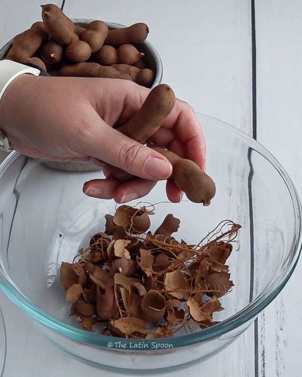 A hand holds two tamarind pods over a glass bowl filled with discarded shells, while another bowl with whole tamarinds sits in the background.