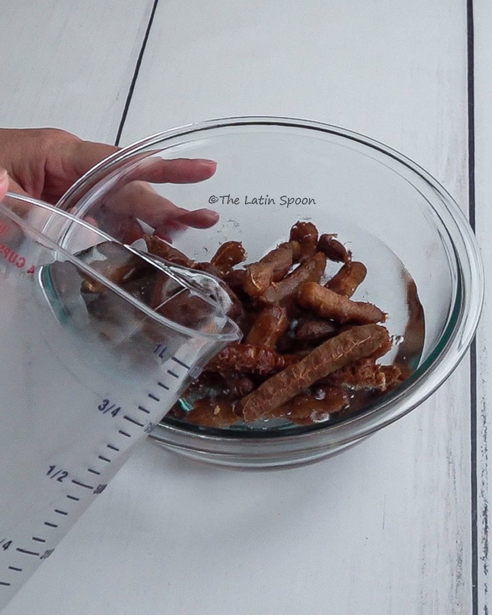 A left hand holds a glass bowl with peeled tamarind, while the right hand tilts a pitcher to pour water into the bowl.