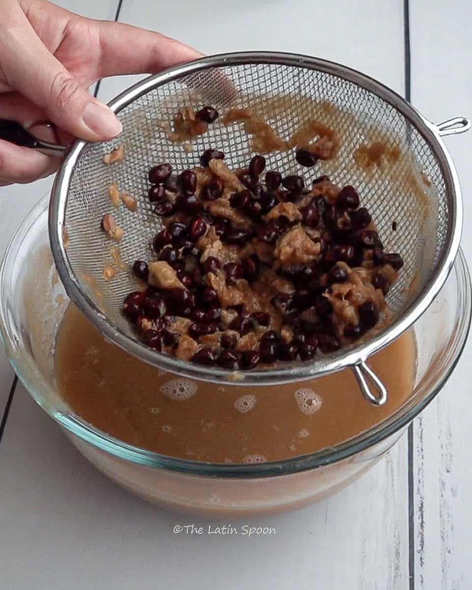 A left hand holds a strainer with tamarind seeds and pulp remains, draining over a bowl.