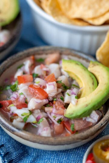 A bowl with small pieces of fish, tomatoes, cilantro, and onion, topped with two slices of avocado. On the side, a small cup with sliced hot peppers and a few tortilla chips.
