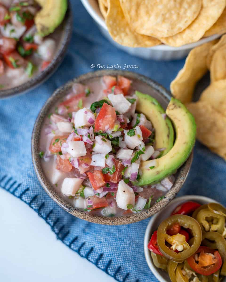 A bowl with small pieces of fish, tomatoes, cilantro, and onion, topped with two slices of avocado. On the side, a small cup with sliced hot peppers and a few tortilla chips.