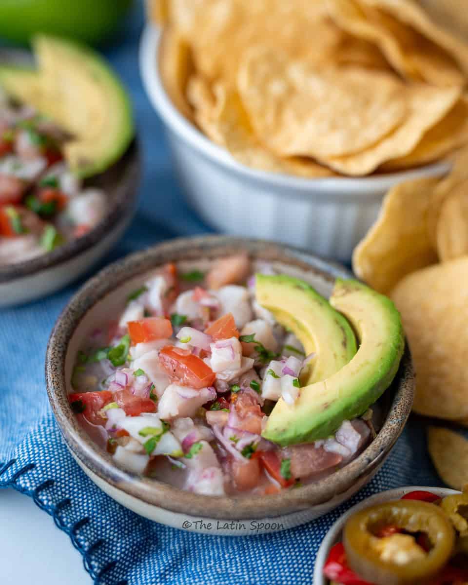 A bowl with small pieces of fish, tomatoes, cilantro, and onion, topped with two slices of avocado. On the side, a small cup with sliced hot peppers and a few tortilla chips.