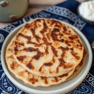 Three cooked Tortillas Aliñadas stacked on a round white plate, placed over a blue cloth, with a bowl of sour cream in the background.