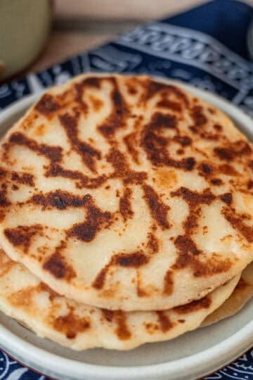 Three cooked Tortillas Aliñadas stacked on a round white plate, placed over a blue cloth, with a bowl of sour cream in the background.