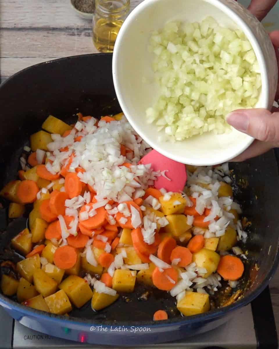 Inside the Dutch oven, you can see potato cubes, carrot slices, and chopped onion, while a hand pours a small cup of celery into the pot. In the background, there’s a container of oil slightly out of focus.