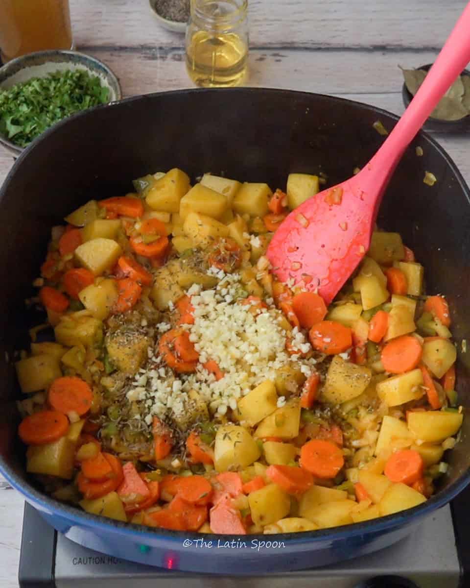 nside the Dutch oven, there are potato cubes, carrot slices, chopped garlic, and oregano, with a spatula resting inside. In the background, a small cup of chopped cilantro, a glass with oil, and a few bay leaves