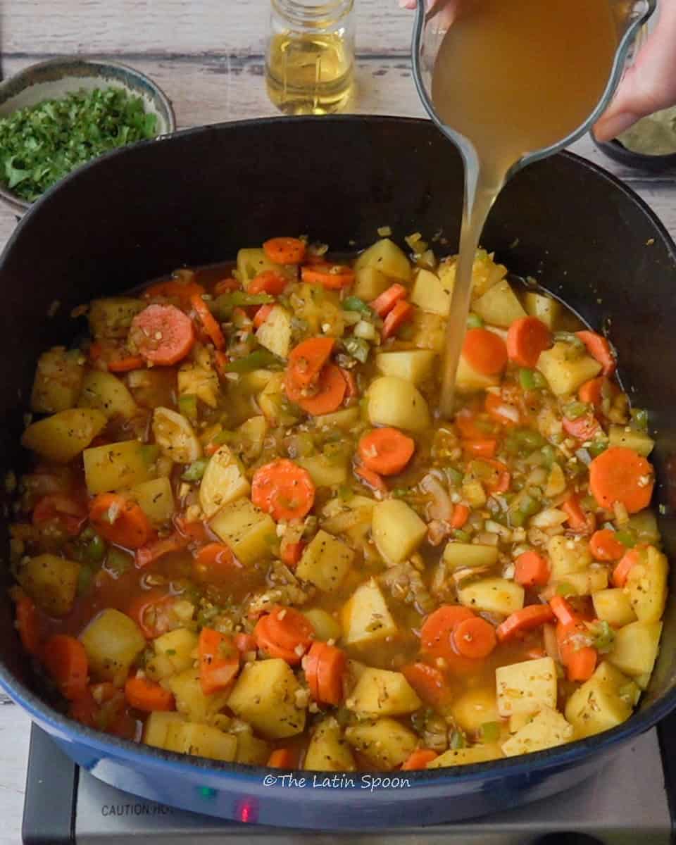 In the pot, there are chunks of carrot and potato mixed with the seasonings, while a container of chicken broth is being poured into the mixture.